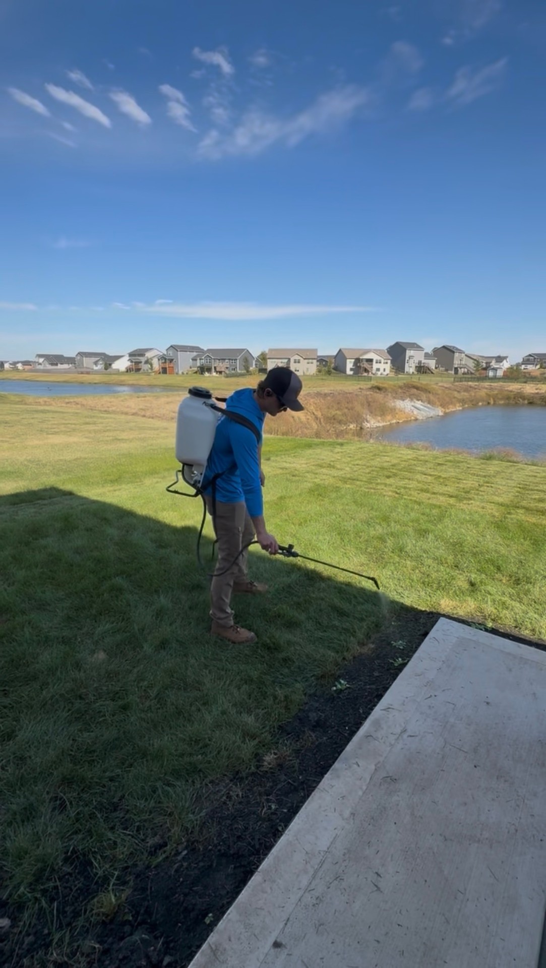 Operator with backpack sprayer applying lawn treatment near a pond in Ankeny